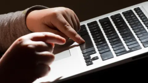 A child's hands typing on a laptop. The laptop's keyboard is black and the child is wearing a long sleeve grey top.
