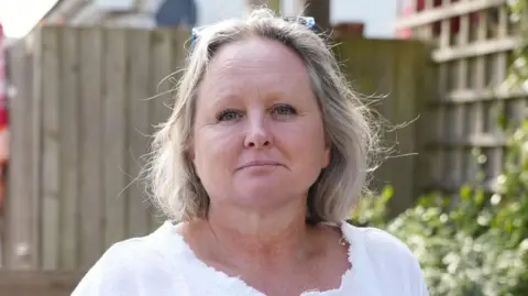 Romy Wood looks at the camera with a neutral expression. She has chin length grey hair and wears a white top, her glasses are perched on the top of her head. Behind her in a wooden garden fence and a trellis with plants on.