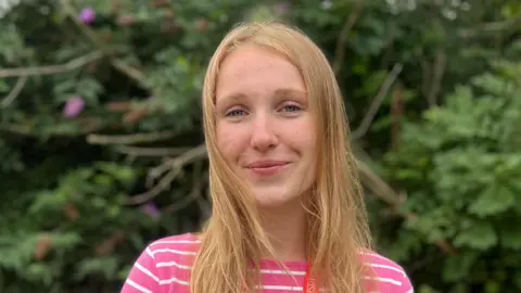 Rosalie Van Zutphen. She has long blonde hair and is wearing a pink and white striped top. She is stood in front of some bushes and flowers.