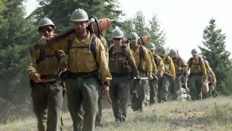 A line of men in firefighting gear walk through an opening in a forest towards the camera lens.