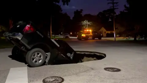 Black pickup truck half submerged in a sinkhole with an emergency vehicle in the background.