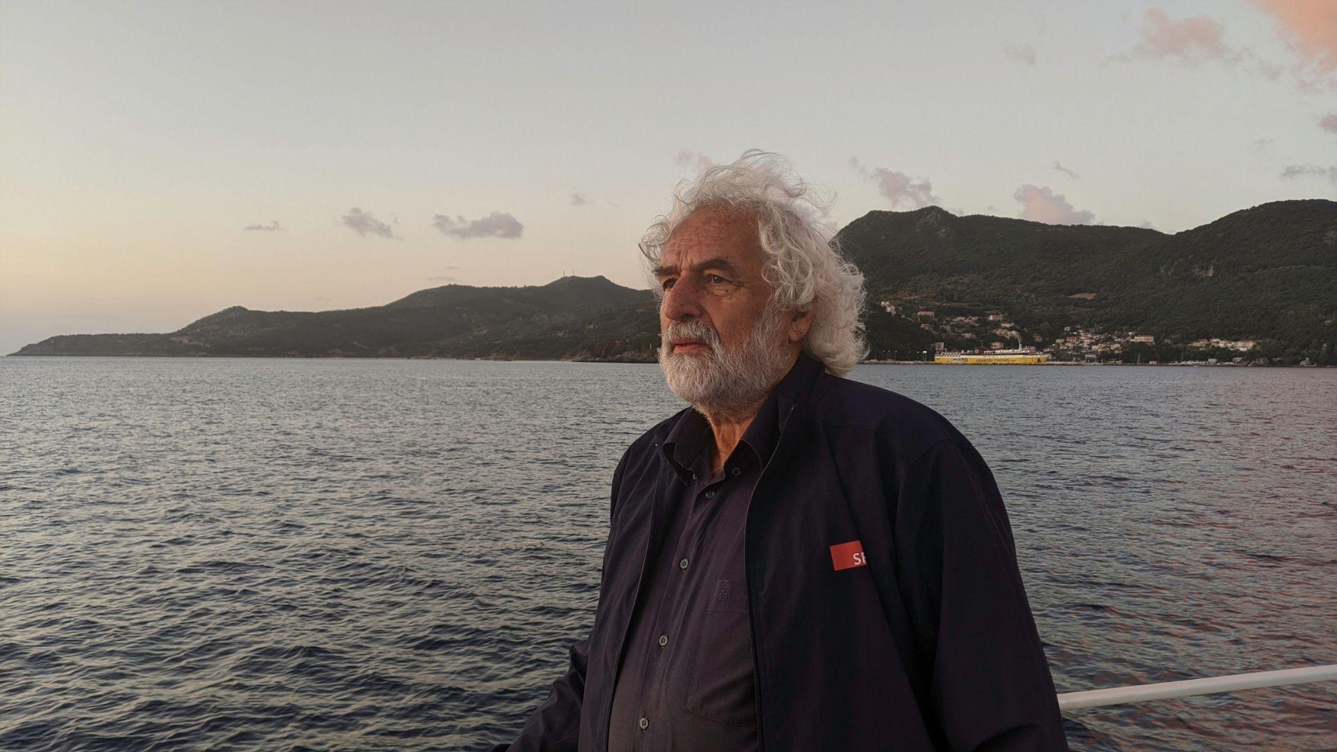 Makis Metaxas, the amateur historian from Kefalonia featured in the documentary Odysseus Returns, standing near the sea with mountainous coastline in the background.