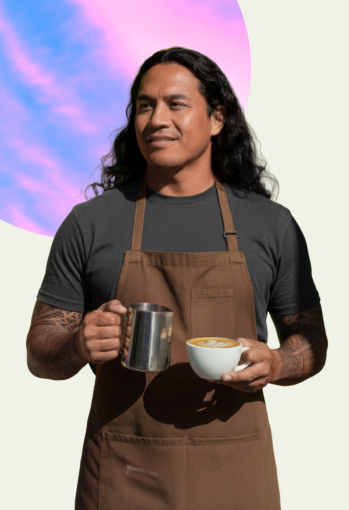 Person operating an espresso machine, steaming milk in a coffee shop. They wear a patterned shirt, jeans, and a beige cap. Framed artwork decorates the minimalist background.