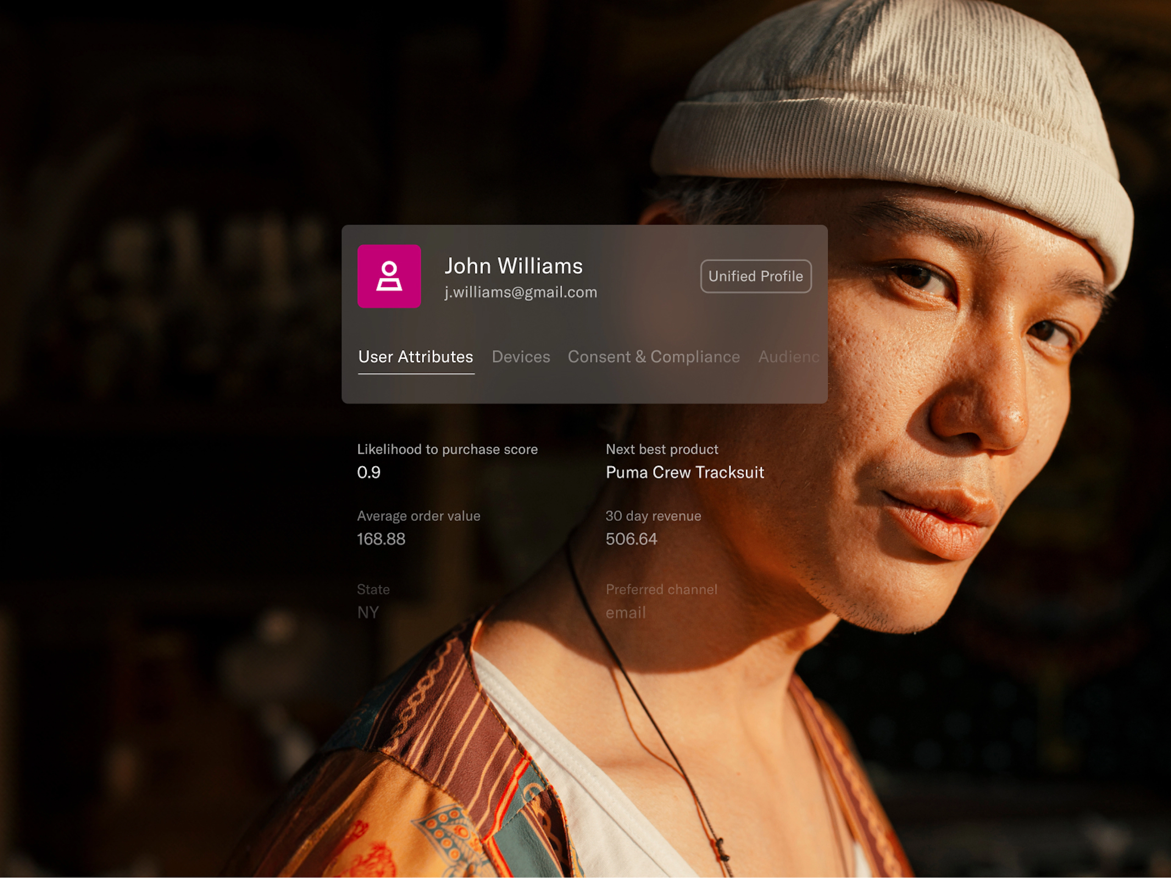 A young man wearing a patterned jacket and a light beige knit cap gazes confidently at the camera in warm sunlight. Overlaid on the image is a digital customer profile card for “John Williams” with an email address and tabs like “User Attributes” and “Devices.” Key metrics shown include a high purchase likelihood score of 0.9, average order value of $168.88, and a suggested next product—Puma Crew Tracksuit. Additional details note a 30-day revenue of $506.64 and preferred communication channel as email. The image illustrates the use of detailed user data to drive relevant marketing recommendations.