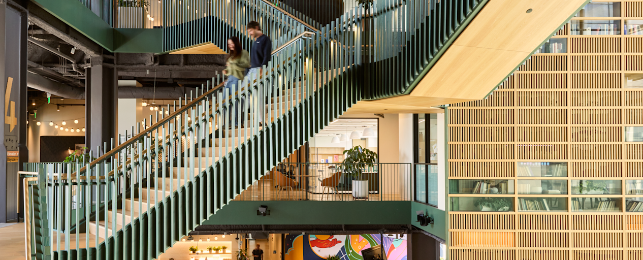 Two people descend a green-railed staircase in an open multi-story office, with a colorful mural on the floor below.