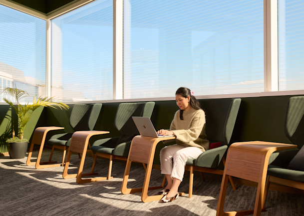 A person working on a laptop in a sunlit office lounge, seated in a row of green chairs along floor-to-ceiling windows.