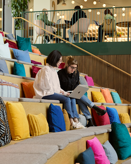 Two people share a laptop on cushion-lined tiered seating, with colleagues working on an open mezzanine above.