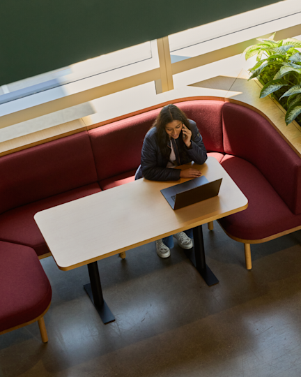 Overhead view of a person on a phone call with a laptop open, seated in a curved red booth beside large windows.
