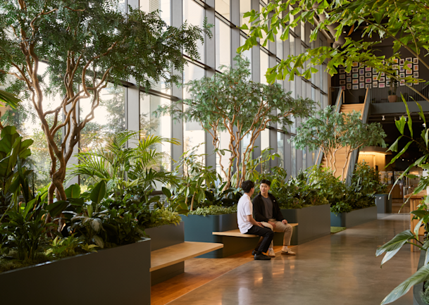 Two people talking on a bench in a sunlit, plant-filled office atrium with floor-to-ceiling windows.