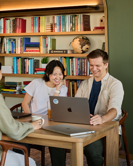 Two people laugh together over laptops at a wooden table in a bookshelf-lined office library.