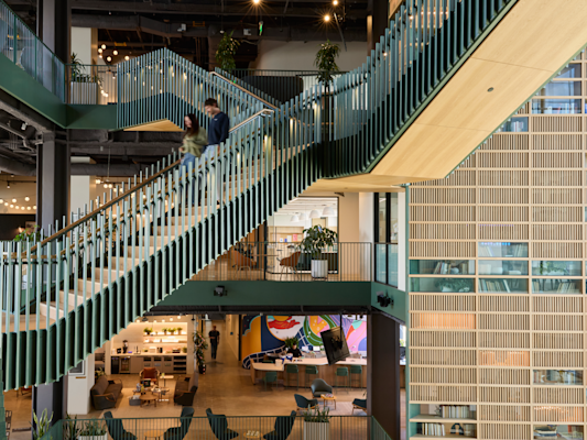 Two people descend a green-railed staircase in an open multi-story office, with a colorful mural on the floor below.