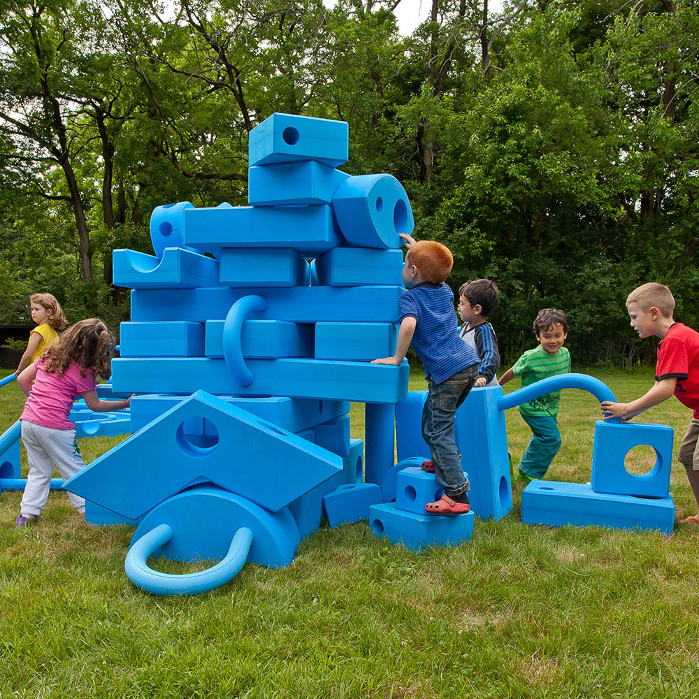 A group of children playing outdoors with Big Blue Blocks, building a tall, complex tower structure on a green lawn. One child is carefully placing a block at the top while others collaborate around the base of the creative foam construction.