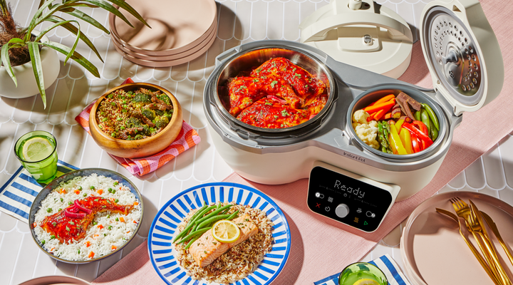 Overhead view of the Sea Salt Dual Action Multi-Cooker open on a table, with a saucy roasted chicken cooking in the larger main pot and colorful vegetables in the smaller side pot. Surrounding the cooker are plated meals including salmon with rice and green beans, chicken and rice, and a grain bowl, along with tableware, drinks, and fresh ingredients.