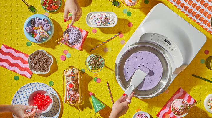 Overhead view of the Sea Salt InstantChill™ Ice Cream Maker in use, with a blackberry ice cream base being spread inside the stainless steel bowl. Surrounding the machine is a colorful spread of toppings and finished treats, including rolled ice cream, scoops with sprinkles, chocolate chunks, cherries, strawberries, and whipped cream on a bright yellow tabletop.