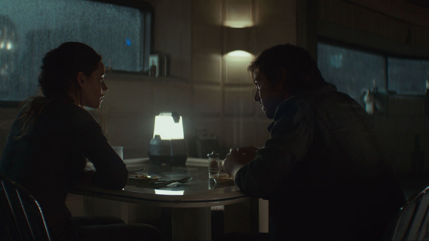 Angled from the back, Bix sits at a table to the left and Cassian sits next to her. The colors are dark and brooding, with a stormy window behind them.