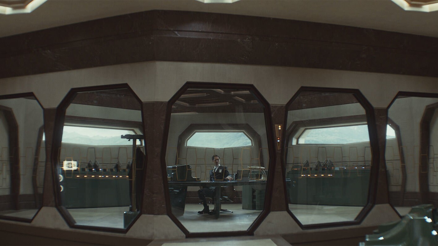 Syril sits in his windowed office. The shot centers him in one of those windows. The interior is brown and dull.
