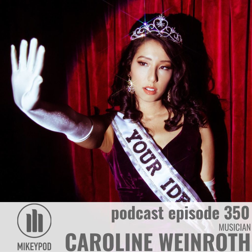 Caroline Weinroth stands in front of a red curtain. Her right hand is extended forward and she is wearing a tiara and a sash with the words "Your Ideal"