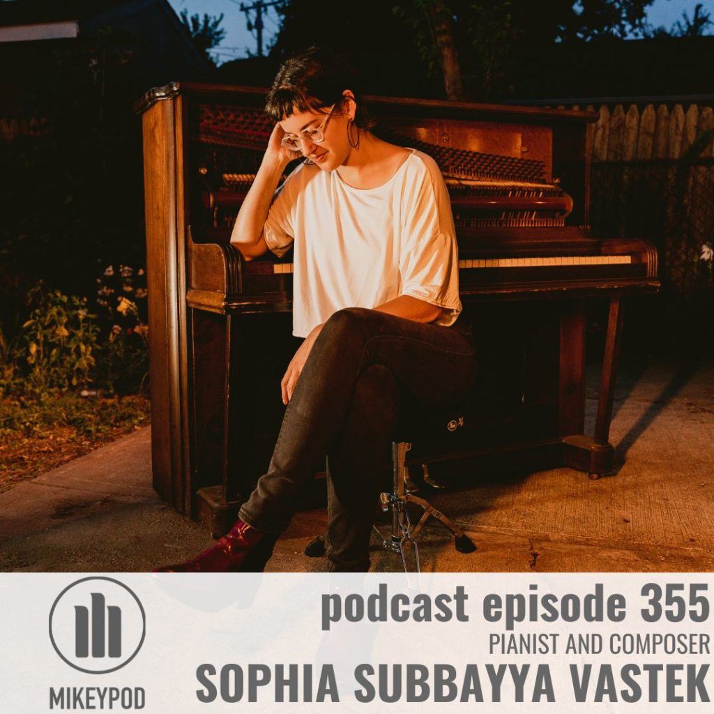 Portrait of Sophia Subbayya Vastek.She is sitting on a stool in front of an upright piano wearing a loose white top, brown boots, and black jeans. Her elbow is resting on the piano.