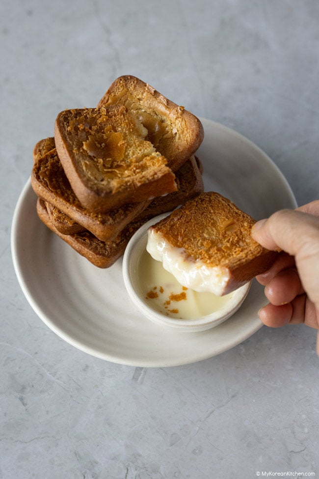 golden butter tteok stacked in the background, with a hand holding a broken piece dipped in sweetened condensed milk