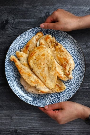 Two hands are holding a blue plate filled with golden brown, perfectly cooked boneless, skinless chicken breasts.