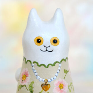 A figurine of a white kitten figurine wearing a pearl necklace with a gold heart locket. The kitten has a tan body with pink flowers and green leaves.