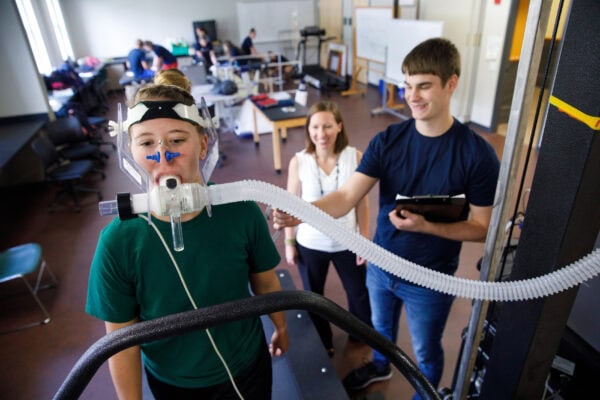 A female student with respiration measuring equipment on sitting on an exercise bike while two other students take measurements.