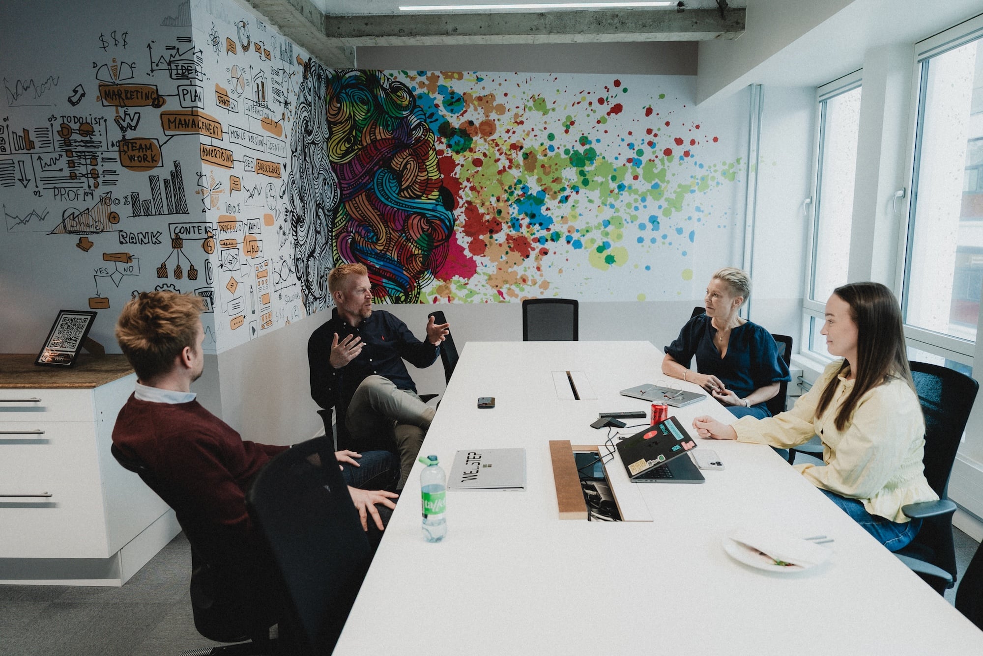 Ansatte i møte rundt et konferansebord på et kontor, med fargerik veggkunst og illustrasjoner som viser idéutvikling, teknologi og samarbeid.