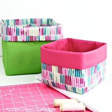two finished handmade fabric baskets in green and pink fabric on a sewing table next to a spool of thread