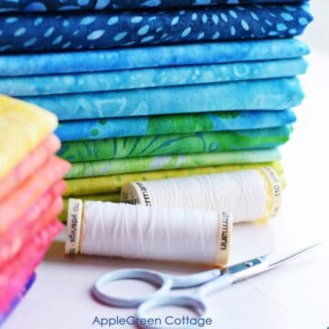 fabric stacked on a table and arranged by color next to small scissors and spools of thread