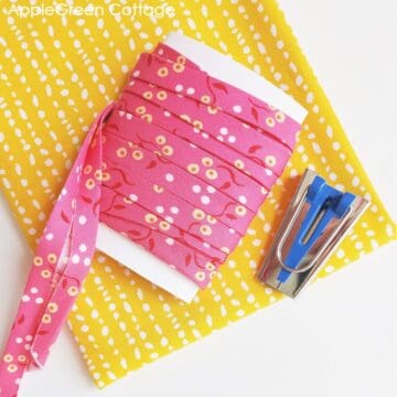 pink double-fold bias tape made from a piece of a liberty print quilting cotton, placed on a yellow print next to a bias tape maker