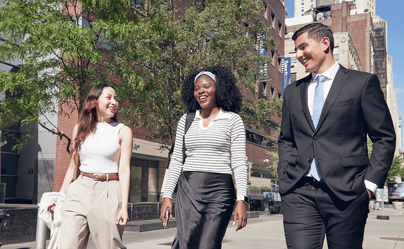 Three students in business-casual attire walking on the Baruch College plaza surrounded by trees and campus buildings.