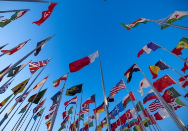 Flags of the world flying on flagpoles against a deep blue sky. Identifiable flags include Bahrain, United Staes, Saudi Arabia, China, France, United Arab Emirates, South Korea, Russia, and the United Kingdom