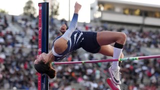 A student athlete in a dark blue and white uniform arches back to clear the bar in the high jump.