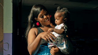 Melissa Robinson, a Black woman wearing a teal blue, magenta and black dress, smiles as she holds her daughter.