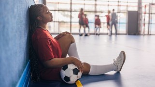 A teen soccer athlete in white socks and a red jersey and sits with a ball against the blue wall of an indoor soccer court. A group of people can be seen in the background.