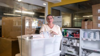 A photograph of a middle-aged Caucasian woman with slicked-back hair sitting in a room with white USPS boxes looking into the camera from behind glass. She leans on a white plastic container nearly overflowing with white carboard USPS boxes.