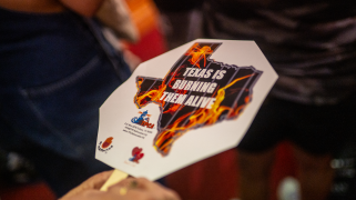 A hand holding a sign with prison cell bars and fire in the shape of the state of Texas. It reads "Texas is burning them alive" in white text.