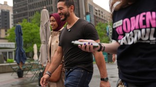 Abdul El-Sayed walks with his wife, Sarah Jukaku, past some picnic chairs and umbrellas in an outdoor city area. Another person holding a phone in their right hand and wearing a "WOMEN FOR ABDUL" shirt is walking to the pair's left.