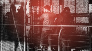 Migrants, seen from behind, stand in a line behind fencing. One man holds his hands to his face, covering his ears. The photo is overlaid with a red-and-black gradient and grainy filter.