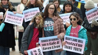 A woman and two children at a trans-rights protest look dejected as they hold signs saying "Gender affirming care is safe and saves lives."