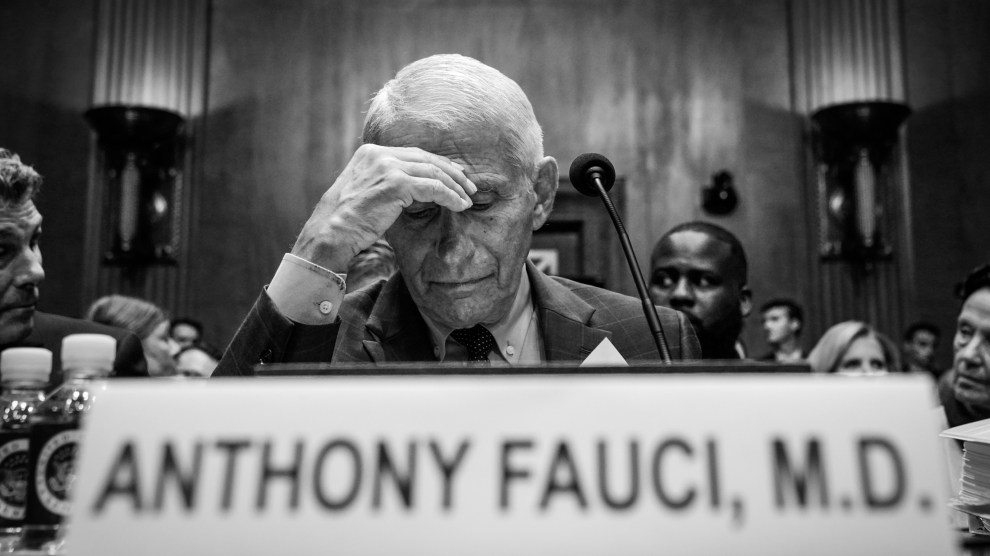An elderly man in a suit with white hair sits at a table behind a microphone and nameplate that reads “Anthony Fauci, M.D.” Appearing agitated, the man is staring downward as he holds his right hand to his forehead.