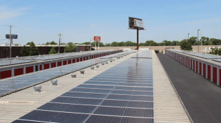 looking along a long row of solar panels on the roof of a storage facility, with rows of identical doors visible below on either side.