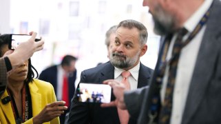 A man with a graying beard and pink tie looks upward as reporters hold out smartphones and microphones around him.