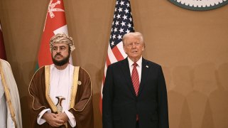 Donald Trump stands next to an official in traditional Omani attire in front of the U.S. and Omani flags.