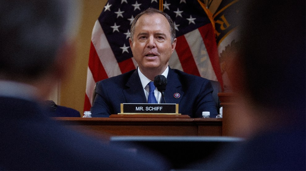 A man in a suit jacket and tie speaks into a microphone. He is seated behind a dais that has a nameplate that reads “Mr. Schiff.” Behind him is the American flag. In front of him, to the left and right, are two men, also in suits, watching Schiff as he speaks.
