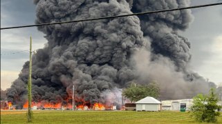 A massive, wide frightening cloud of dark smoke rises from a burning industrial facility