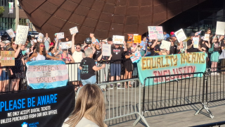 An individual wearing a black shirt and blue pants walks down a line of rallygoers hold signs supporting trans rights and inclusion in sports.