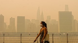 A woman walks through a smoke-filled park wearing a mask, with the skyline of New York City in the background through the haze.