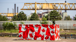 Dozens of Canadian flags cover a utilities box outside a steel plant. In the background is yellow construction scaffolding, a street light, a power line, and factory-looking structures.