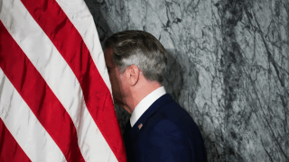 Scott Bessent is walking to the left. His face is behind an American flag and in shadow, but the photo shows his hair, neck, and shoulders from a side profile. He is walking in front of a gray wall.